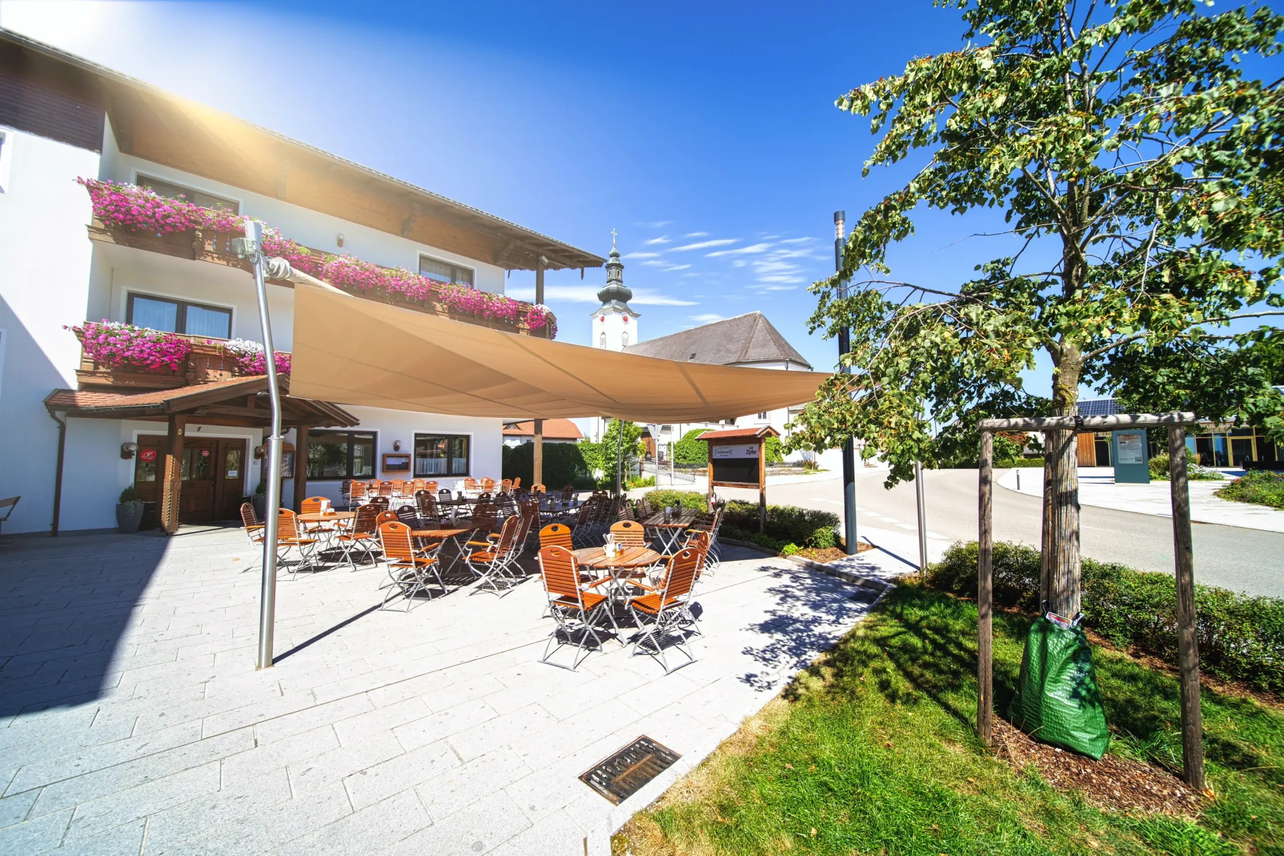 Modernes Sonnensegel beim Kirchenwirt St. Aegidi mit Blick auf die Kirche; textile Architektur schützt den öffentlichen Versammlungsplatz.