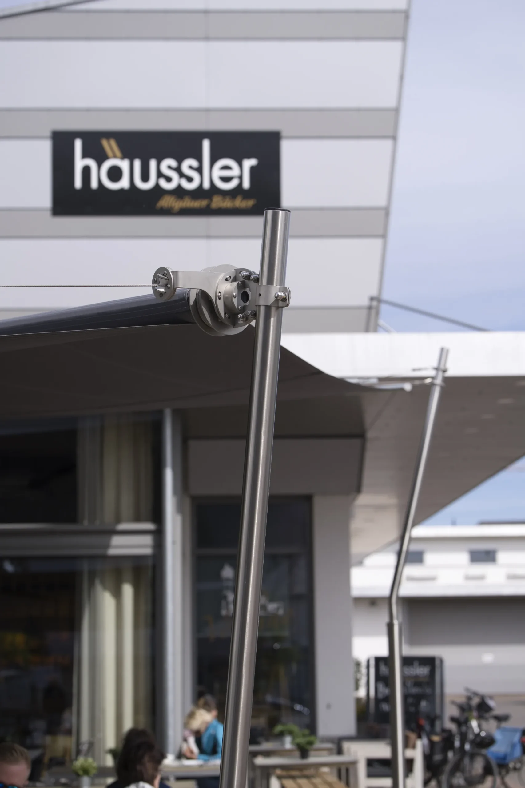 Edelstahlmast des Sonnensegels vor dem Logo von Backhaus Häussler an der Fassade in Illertissen