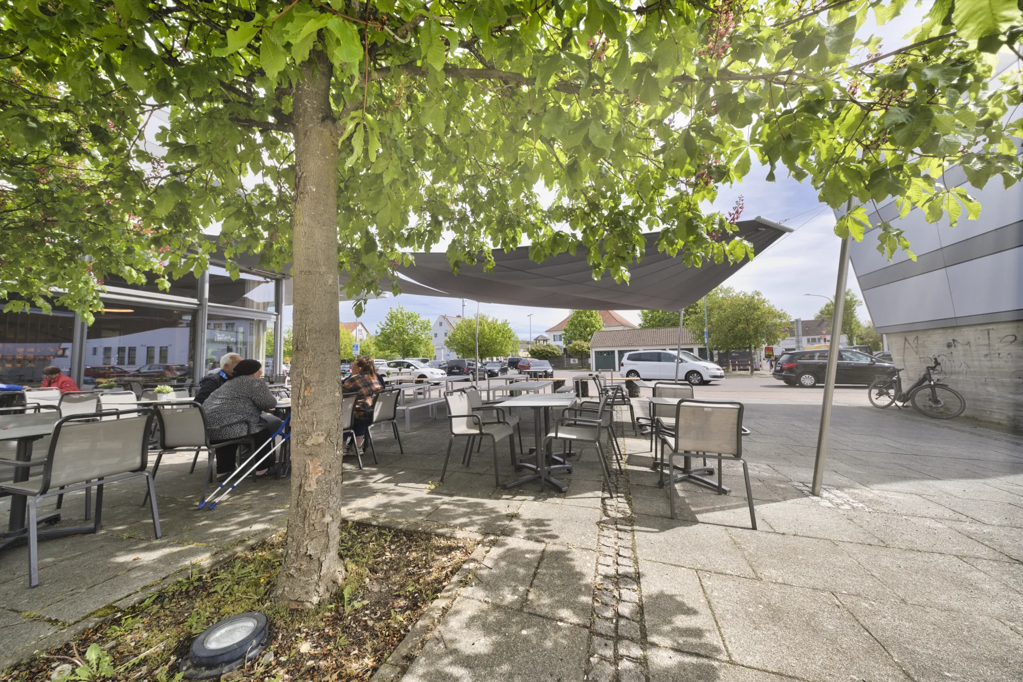 Überblick der Café-Terrasse von Backhaus Häussler unter dem Sonnensegel mit Gästen, Kastanie und Sitzplätzen in Illertissen
